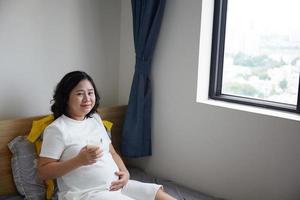 Happy Young Asian Pregnant Woman drinking milk on bed at home while holding her belly and thinking photo