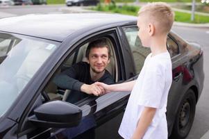 un linda niño batidos manos con un contento padre . papá es sentado en el coche y sonriente foto
