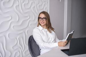 A young teacher with glasses holds a diary in her hands and sits at a table with a laptop photo