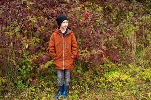 A cute boy in rubber boots stands in the autumn forest photo