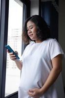 Young Asian Pregnant Woman standing near the window and using smart phone for checking information photo