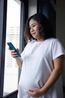Young Asian Pregnant Woman standing near the window and using smart phone for checking information photo