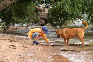 A boy in an orange shirt plays with a red dog on the seashore. photo