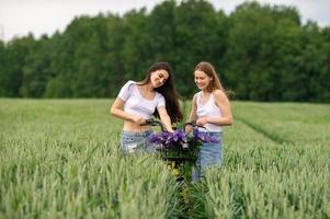 Two pretty girls are standing with a bouquet of lupines in a wheat field next to a bicycle photo