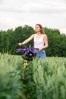 un linda niña es en pie con un ramo de flores de altramuces en un campo cerca un bicicleta foto