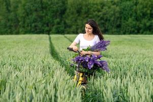 un bonito niña soportes con un ramo de flores de altramuces en un campo siguiente a un bicicleta foto