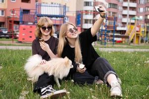 dos gracioso muchachas son comiendo hielo crema y jugando con un pomeranio perro. tomando selfies foto