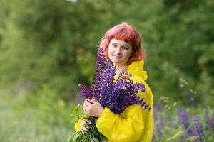 A cute teenage girl with pink hair and a bouquet of lupines is standing in a yellow raincoat photo
