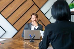 A man in a suit is sitting with a laptop in the office and asking questions to the interlocutor photo