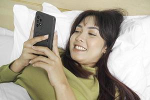 A smiling young Asian woman is laughing while holding her phone and lying on the bed photo