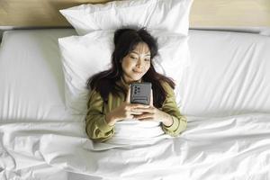 A smiling young Asian woman is laughing while holding her phone and lying on the bed photo