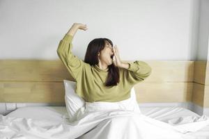 Cute Asian woman stretching out and yawning while sitting on her bed in bright bedroom photo