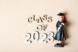 Class of 2023 concept. Wooden number 2023 with graduate statuette on beige background photo