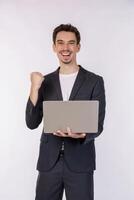 Portrait of young smiling businessman holding laptop in hands, typing and browsing web pages while doing a winning closed fist gesture isolated on white background photo