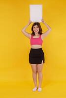 Portrait of Happy young woman holding an empty white placard over isolated yellow background. photo