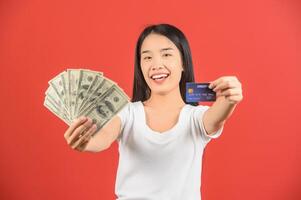 Portrait of a cheerful young woman holding money banknotes and credit card isolated over red background photo