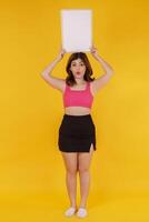 Portrait of Happy young woman holding an empty white placard over isolated yellow background. photo