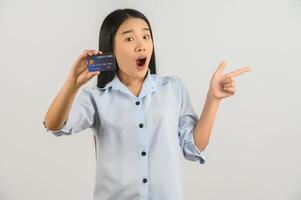 Portrait of positive young asian woman showing credit card good mood salary and pointing finger at aside isolated on white background photo