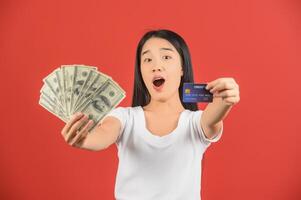 Portrait of a cheerful young woman holding money banknotes and credit card isolated over red background photo