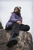 retrato de un linda de moda caucásico niña vestido en un calentar de punto sombrero, púrpura abajo Saco sentado en un desnudo Roca foto