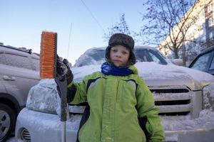 Funny cute Caucasian boy cleans the car from snow with a brush and a scraper photo