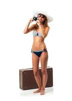 Full length portrait of a beautiful young woman posing in a bikini with a camera in hands and suitcase on white photo