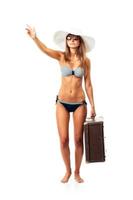 Full length portrait of a beautiful young woman posing in a bikini, hat and sunglasses with a suitcase in hand on white photo