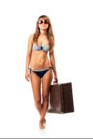 lleno longitud retrato de un hermosa joven mujer posando en un bikini y Gafas de sol con un maleta en mano en blanco foto