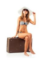 Full length portrait of a beautiful young woman posing in a bikini sitting on a suitcase on a white photo