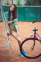 hermosa joven niña con longboard y bicicleta en pie en el tenis Corte foto