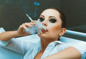 joven niña en un blanco camisa con brillante maquillaje acostado en el baño y fuma un cigarrillo foto