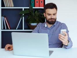 Bearded man working on a laptop in the office and talking on cell phone photo