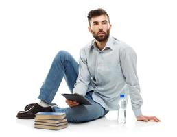 Young bearded man holding a tablet with books and a bottle of water sitting on a white photo