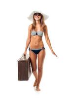 Full length portrait of a beautiful young woman posing in a bikini, hat and sunglasses with a suitcase in hand on white photo