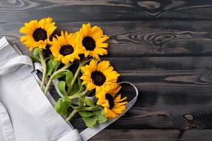 fresh sunflowers with leaves on stalk in shopping bag on wooden background. Flat lay, top view, copy space. photo