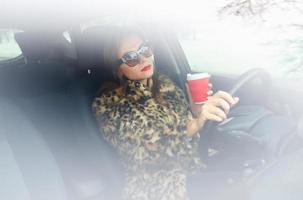Woman in a fur coat with red lips with coffee to go driving her car photo