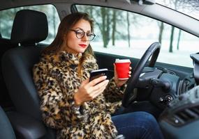 mujer de negocios en un piel Saco con rojo labios enviando un texto mensaje y Bebiendo café mientras conducción foto