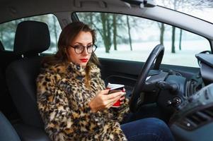 Businesswoman in a fur coat with red lips sending a text message and drinking coffee while driving photo