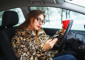 Businesswoman in a fur coat with red lips sending a text message and drinking coffee while driving photo