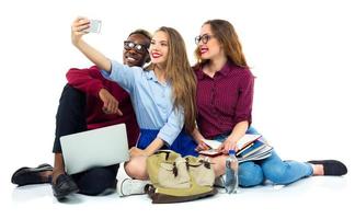 Tres contento estudiantes con libros, computadora portátil, pantalones y hace selfie foto