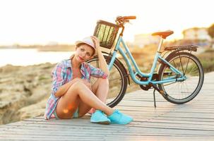 despreocupado mujer con bicicleta sentado en un de madera camino a el mar foto