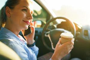 Young woman talking on a smartphone and drinking coffee while driving a car photo