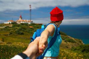 Follow me - happy young woman in a red hat and with a backpack behind her back pulling guy's hand. Hand in hand walking to the lighthouse photo