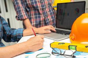 Asian engineer and partner team learn and talk with house plan project blueprint, graph and yellow helmet and engineering construction tools on table work. photo