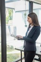 A stylish white businesswoman in a black suit standing inside the office in hands with a mobile phone, tablet, and laptop waiting for customers to contact, business people concept. photo
