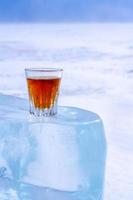 Whiskey in a glass stands on an ice block. Orange alcoholic drink in a glass. The ice melted in the sun. Light blurred background with snow. Copy space. Side view. Vertical. photo