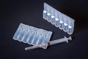 un desechable jeringuilla para personal utilizar y dos paquetes de el plastico ampollas en un oscuro antecedentes. líquido en ampollas viñeteado horizontal. foto