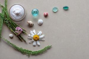 Flat view. Still life for spa. Green spikelets and plants, purple flowers, white with yellow daisy, colorful shells, glass and a jar of white cream. Selective focus. Background brown ceramic tile. photo