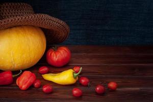 Pumpkin in a hat, scattered red tomatoes and peppers, one yellow pepper. Tomatoes are small and one big. Boards are brown, a background from a scarf fabric. Suitable for halloween. Vignette. photo