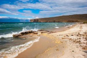 The shore of Lake Baikal with splashes from the waves breaking on stones. Sandy shore. A rocky cape in the distance. Clouds in the sky. Horizontal. photo